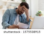 Small photo of Upset frustrated young man reading bad news in postal mail letter paper document sit at home table, depressed stressed guy worried about high bill tax invoice, overdue debt notification money problem