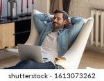 Peaceful young man daydreaming in comfortable armchair with computer on knees. Calm guy taking break after working with laptop, controlling fatigue or stress, resting alone in living room at home.