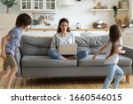 Small photo of Calm young mother or nanny sit on couch working on laptop, little kids play around, peaceful mom relax on sofa use modern computer distracted from noise, parenting, upbringing concept