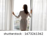 Rear back view young happy brunette woman in white night gown standing near opened curtains huge panoramic window, looking outside, enjoying view at hotel home, good morning lifestyle concept.