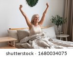 Small photo of Joyful healthy older woman sitting on bed, waking up after good night rest on comfortable orthopedic mattress, rising up arms and stretching back. Happy mature grandma feeling energetic in morning.
