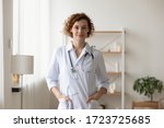 Confident young female physician standing in medical office. Proud professional woman doctor therapist looking at camera. Portrait of lady general practitioner wearing white coat and stethoscope.