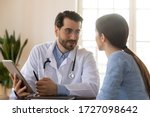 Small photo of Smiling young Caucasian male doctor in white medical uniform consult female patient using tablet on consultation, modern man GP talk discuss checkup results with woman client at meeting in hospital