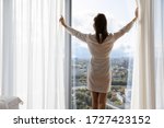 Rested female in white night robe wake up opens curtains enjoys big city and sunny weather, rear view. Welcoming of new day, good morning, modern warm hotel bedroom, luxury apartments owner concept