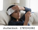 Small photo of Sick millennial Asian sit on couch at home with towel on head measure temperate on thermometer. Unhealthy ill young Vietnamese woman suffer from flu or grippe, catch cold. Health problem concept