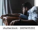 Upset young African American male sit on sofa at home look in distance mourning missing. Unhappy sad biracial guy distressed with life problems, feel lonely thinking or pondering. Solitude concept.