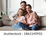 Loving young man hug his beloved wife and little daughter sit on couch in living room, happy people smiling looking at camera posing for photo picture. Exemplary family portrait, love and bond concept