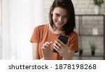 Small photo of Calling to someone especial. Happy millennial woman stand at living room hold phone in hands dial number on touchscreen. Cheerful young lady surfing internet choosing name from contact list on cell