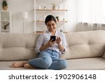 Small photo of Happy indian lady relax at home alone sit on large couch in comfortable pose share good news at social media via cellphone. Smiling mixed race woman enjoy weekend order goods food online in phone app