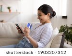 Small photo of Young indian woman shopper sit on couch enjoy easy paying for goods online using bank card phone. Happy mixed race female traveler book tickets trip hotel at travel agency transport operator website