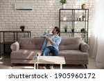 Full length young man wearing glasses holding using air conditioner remote controller, relaxing, sitting on couch, switching, setting comfort temperature in modern living room, enjoy fresh air