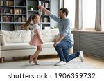Small photo of Full length happy young daddy standing on one knee, twisting smiling adorable preschool kid daughter in princess dress, practicing waltz or preparing performance, dancing together in living room.