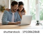 Small photo of Cheering spouses having warm relations hug by desk at home make shopping purchases online at web store. Friendly young couple in love look at computer screen planning wedding choosing tour on vacation