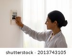 Close up smiling Indian using smart home system controller on wall, positive attractive young female switching temperature on thermostat or activating or turning off security alarm in apartment