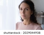 Small photo of Close up portrait of joyless 30s woman standing alone near window indoor. Face of Hispanic serious female with sad eyes staring at camera. Tiredness, lack of optimism, solitude, life concerns concept