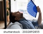 Small photo of Exhausted overheated millennial man suffering from heat at home without conditioner, trying to cool too hot air with handheld fan, feeling unwell due to weather, humidity, air temperature, hypoxia