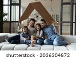 Happy sibling kids and parents holding carton toy roof above heads, showing symbol of home, safety, celebrating buying new house, property, health insurance. Real estate, mortgage, family concept