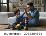 Happy millennial 30s dad giving praise and high five to little son for building toy tower from construction plastic blocks. Joyful kid playing learning games with daddy on heating floor