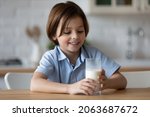 Small photo of Happy boy with funny white moustache drinking milk in kitchen, holding glass, smiling. Cheerful kid keeping healthy diet nutrition, getting calcium, vitamins, probiotics for growth from dairy products