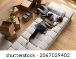 Overhead view peaceful young couple relaxing on fashionable cozy sofa near heap of packed belongings in cardboard boxes, resting at own house at relocation day. Bank loan and mortgage, tenancy concept