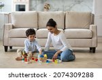 Small photo of Happy young Indian mom helping preschool son to build toy towers from wooden construction blocks, playing with kid on heating floor at home. Babysitter watching child. Motherhood, daycare concept