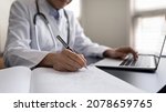 Small photo of Crop close up Indian woman doctor in white uniform with stethoscope taking notes, using laptop, writing in medical journal, professional therapist practitioner filling documents or patient card