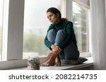 Heavy heart. Sad hispanic lady sit barefoot on windowsill feel absent minded tired stressed with problems in relations. Depressed young female look down the window try to overcome psychological trauma