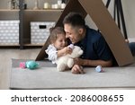 Small photo of Happy playful daughter kid and loving daddy playing tea party in toy handmade house, relaxing on warm floor under cardboard paper roof, using childish colorful plastic dish. Childhood, housing