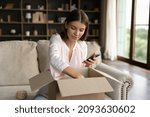 Young woman, shopper, consumer receiving parcel, unboxing package from internet store, producing goods from cardboard box, using shopping app on mobile phone, giving feedback to order delivery service