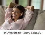 Cheerful relaxed young woman with wireless earbud resting on sofa, listening to ambient tunes, positive musing for relaxation. Woman using earphones, enjoying weekend, leisure time at home