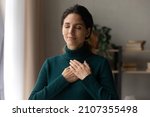 Head shot peaceful young attractive woman holding folded hands on chest, praying waiting for miracle, feeling thankful indoors. Sincere happy female volunteer showing kindness or expressing gratitude.