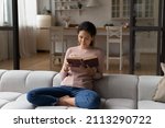 Small photo of Young 30s Hispanic woman holding paper book seated cross-legged on sofa at home, reading exciting bestseller literature, enjoy lazy weekend, day off free time with favourite hobby. Leisure concept