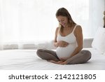 Small photo of Happy expectant mother sitting on bed, feeling kicks, caressing big belly, smiling. Pregnant woman in white tank top touching bump, speaking to unborn baby. Motherhood, pregnancy concept
