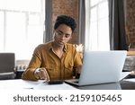 Small photo of Serious African woman sit at desk with laptop, analyzing bills, do accounting job, review utilities, calculates summary for payment through electronic bank app. Finance management, bookkeeping concept
