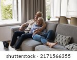 Joyful older grey-haired couple in love hugging relaxing on sofa in living room laughing while watching comedy movie on TV. Happy carefree spouses talking feel overjoyed spend weekend leisure at home