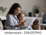 Small photo of Loving mother reading a book to her 6s daughter, family sit on cozy sofa covered with warm knitted plaid enjoy favourite hobby. Children development, fascinating bestseller story, bookshop ad concept