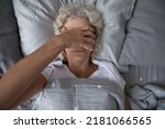 Small photo of Peaceful sleepy senior woman awaking in her bed, rubbing face, covering eyes with hand from disturbing light. Elderly mature lady suffering from insomnia, sleep disorder or deprivation. Top view