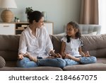 Cheerful Indian mom teaching girl to do yoga at home, sitting in lotus pose on couch, talking to daughter kid, laughing, smiling, discussing meditation, enjoying activity, exercises, family leisure