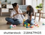 Small photo of Happy beautiful Indian mom and pretty little daughter girl clapping hands, giving high five over wooden construction blocks, toy tower, castle on heating floor, celebrating model completing