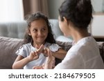 Small photo of Happy cute Indian kid girl talking to mother, using hands, fingers, speaking sign languages, sitting on sofa at home. Female teacher, therapist teaching child with deafness to communicate