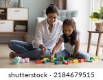 Small photo of Cheerful pretty Indian mother and little daughter kid playing on heating floor at home, arranging colorful toy blocks. Nanny, daycare teacher entertaining child, enjoying learning game