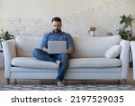 Handsome serious man sits on sofa with computer on laps, spend time at home web surfing information, use modern tech, make order on internet. Freelancing, telework, new software learning, tech concept