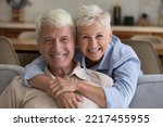 Small photo of Grey-haired grandparents pose at home. Loving wife hugs husband seated on sofa staring at camera, spend time together. Happy long-lasting marriage, harmonic relations between spouses, respect and bond