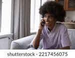 Small photo of Curly-haired African teenager guy sits on sofa blab on smart phone, holds mobile phone enjoy pleasant conversation, communicates remotely with girlfriend using modern tech, enjoy personal phonecall