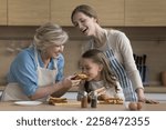 Small photo of Cheerful little girl, mom and grandma enjoying culinary activity in kitchen, cooking fast food snacks for lunch. Happy grandmother giving sandwich for kid biting, taste, feeding hungry child