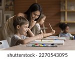 Small photo of Cheerful pretty teacher girl helping pupil boy with drawing picture on canvas, standing at kid, smiling, laughing, looking at canvas. Child attending artistic school class, learning painting
