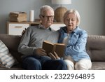 Small photo of Focused mature spouses reading a book together at home. Serious baby-boomer generation spouses spend pastime resting on couch at home, enjoy favourite hobby, bestseller readers, literature store ad