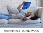 Small photo of Exhausted sick homeowner woman resting on home sofa, waving handheld paper fan, suffering from fatigue, hypoxia, headache, heating stroke, holding head, swiping sweat