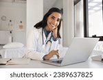 Small photo of Cheerful attractive doctor woman professional portrait. Young Latin practitioner sitting at workplace table with laptop, looking at camera with toothy smile posing for shooting in surgery office