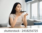 Small photo of Serious young adult Chinese smartphone user girl talking on speaker, speaking at mobile phone, recording voice audio message, using devise for wireless online communication